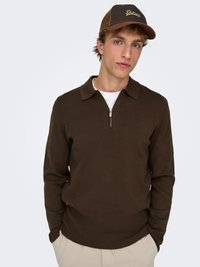 Maglione marrone lavorato a maglia con colletto con zip, abbinato a un cappellino marrone e in rete con la scritta "Detroit" in corsivo. Il modello è in piedi con le mani nelle tasche.
