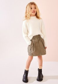 Weißer gerippter Strickpullover, olivgrüner ausgestellter Rock mit seitlichen Taschen, schwarze Ankle Boots mit klobigen Sohlen und cremefarbene Socken.