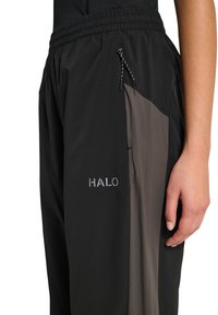 Nahaufnahme von schwarzen und grauen Sporthosen mit elastischem Bund, seitlichem Kordelzug und "HALO"-Logo am Oberschenkel, neben denen ein Arm einer Person ruht.