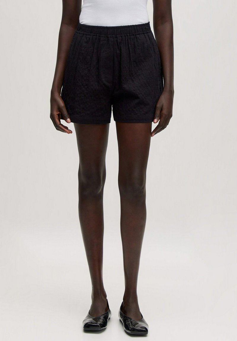 Zwarte, gestructureerde shorts met een elastische tailleband, voorzien van zijzakken en een ontspannen pasvorm. Draagt met een eenvoudige witte tanktop en flats.