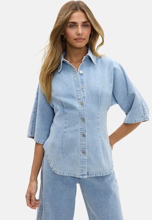 Femme aux longs cheveux blonds, portant une chemise en jean bleu clair à boutons avec de larges manches trois-quarts et un pantalon en jean assorti.