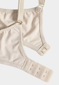Soutien-gorge beige en tissu doux, avec bretelles réglables et fermeture à crochets à trois positions. Texture lisse et coutures minimales.