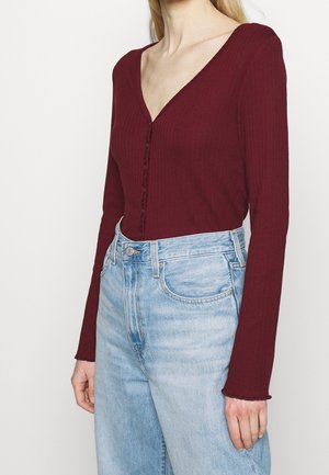 Woman wearing a fitted burgundy ribbed long-sleeve top with buttons paired with light blue high-waisted jeans against a plain background.
