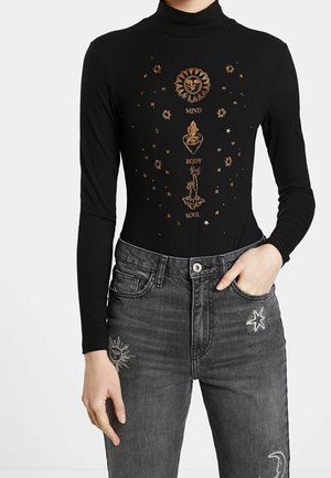 Zwarte coltrui met gouden hemelse ontwerpen en de tekst "Mind Body Soul", gecombineerd met grijze jeans met borduursels van zon, ster en maan.