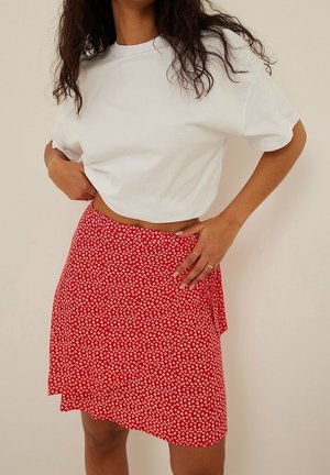 Mujer con cabello largo y rizado, vistiendo una camiseta blanca corta y una falda envolvente roja con flores, con las manos apoyadas en la cintura y la cadera.