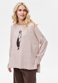 Jasnoróżowy sweter z długimi rękawami, z grafiką kobiety na przodzie; miękka faktura; w połączeniu z brązowymi sztruksowymi spodniami.