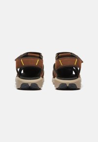 Ruske sandaalit avoimilla takapuoilla, säädettävillä hihnoilla ja keltaisilla yksityiskohdilla. Rihkan musta pohja ja beiget, kestävät pohjat, jotka on suunniteltu pidon parantamiseksi.