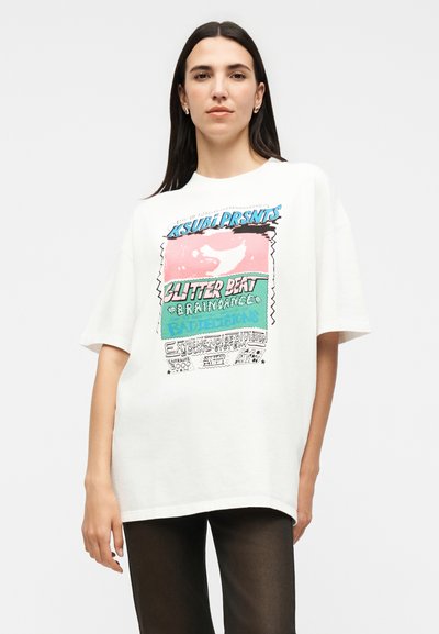 Γυναίκα με μακριά σκούρα μαλλιά που φοράει oversized λευκό μπλουζάκι με πολύχρωμο κείμενο και σχέδιο, συνδυασμένο με μαύρο παντελόνι, στέκεται μπροστά σε λευκό φόντο.