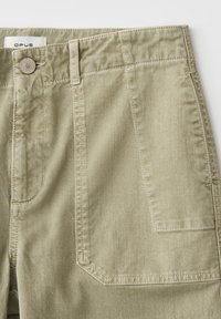 Pantalon en denim beige avec poche plaquée à l'avant, passants pour ceinture, bouton en métal gravé "OPUS" et une étiquette blanche portant le nom de la marque à l'intérieur de la ceinture.