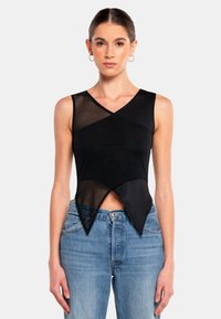 Svart ärmlös topp med en meshsektion, överlappande design och asymmetrisk fåll. Kombinerad med ljusblå jeans.