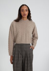 Beiger Strickpullover mit Kabelmuster, hohem Kragen und verkürzter Silhouette, kombiniert mit karrierten, weit geschnittenen Hosen mit Vorderfalten und Taschen.