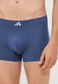 Temno modri bokseriči, izdelani iz raztegljivega materiala, z mehkimi pasom in svetlo sivo Adidasovo logo na sprednji strani.