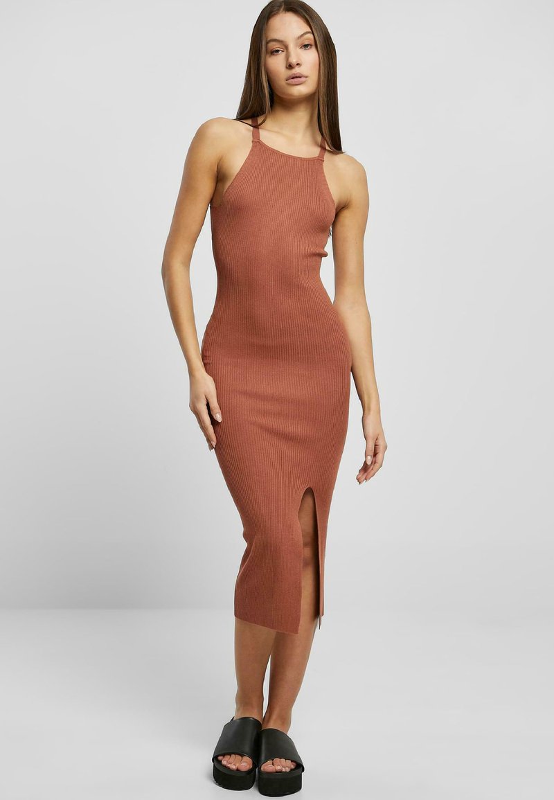 Rippendes, ärmelloses Midikleid in Terrakotta mit einem Seitenschlitz, das eine figurbetonte Silhouette und einen runden Ausschnitt aufweist. Kombiniert mit schwarzen Schuppensandalen.