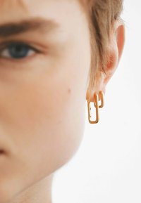 Pendientes de metal dorado con un diseño geométrico de clip de papel, dos lazos interconectados con un acabado suave.