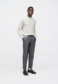 Gaiši pelēks svītrains džemperis ar apaļu apkakli, kombinēts ar tumšām svītrainām biksēm. Melni apavi ar baltiem akcentiem. Vienkārša, moderna silueta.