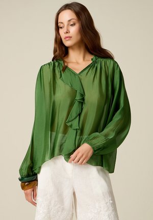 Kvinde iført gennemsigtig grøn bluse med flæser og hvide broderede shorts, poserer mod en ensfarvet beige baggrund.