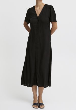 Femme portant une robe midi noire avec des manches courtes bouffantes, une patte de boutonnage sur le devant et un décolleté en V profond, associée à des mules noires à bouts ouverts.