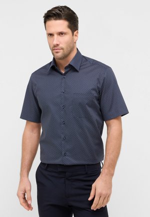 Homme portant une chemise ajustée bleu marine à manches courtes avec un motif de petits points rouges et blancs, associée à un pantalon foncé.