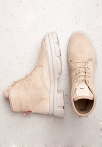 Botas de ante beige con material texturizado, forma estructurada y punta redonda. Presentan una suela blanca con herrajes visibles y acentos rojos.