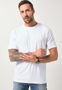 Camiseta blanca de manga corta hecha de algodón, con cuello redondo, textura suave, combinada con jeans azules, mostrando tatuajes en los brazos y un reloj.