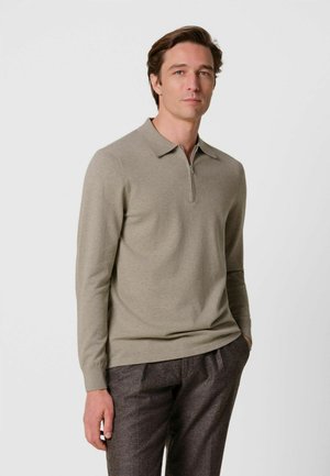 Uomo che indossa una polo beige a maniche lunghe con zip e pantaloni grigio scuro, in piedi con una mano in tasca davanti a uno sfondo semplice.