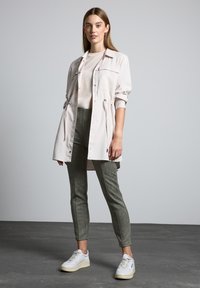 Ljusbeige skjortjacka med dragsko i midjan, ihop med olivgröna tajta byxor och vita sneakers. Mot en neutral bakgrund.