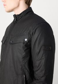 Schwarze Jacke aus Nylon mit hohem Kragen, Reißverschluss vorne, zwei Brusttaschen und einem Abzeichen am Ärmel. Glatte Textur, moderner Schnitt.