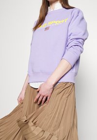 Lila sweatshirt med gul "POLO SPORT" text och en amerikansk flagga patch, kombinerad med en plisserad beige kjol. Mjuk tygkvalitet, avslappnad passform.