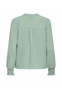Blouse vert clair avec une surface texturée, manches longues et poignets froncés. Elle présente un dos droit et un détail de pli subtil.