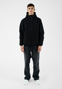 Giacca nera con cappuccio, chiusura con zip frontale, polsini elasticizzati e tasche laterali, abbinata a jeans scuri e sneaker chiare su uno sfondo neutro.