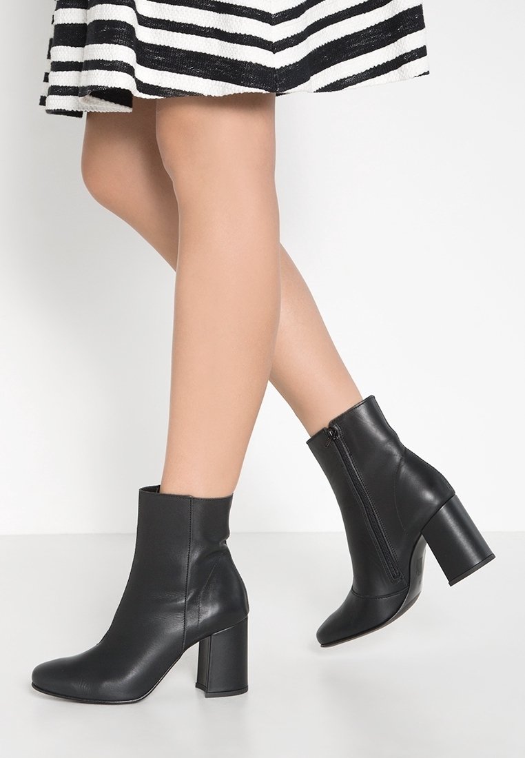 Jambes portant des bottines en cuir noir à talons blocs, associées à une jupe rayée noire et blanche sur un fond blanc uni.