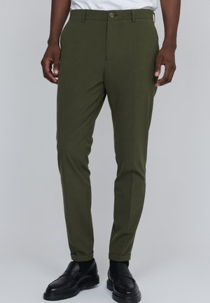 Uomo che indossa pantaloni verde oliva slim-fit, calzini neri e mocassini neri in pelle, in piedi davanti a uno sfondo neutro.