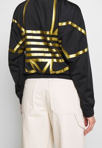 Black bomber jacket with gold stripe pattern and detailing. Features elastic cuffs and hem, with a slightly loose fit. Light, smooth material.