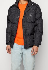 Czarna kurtka puchowa z tłoczonymi geometrycznymi wzorami, zapinana na zamek błyskawiczny, z kołnierzem na zatrzaski. Noszona na pomarańczowej koszuli, w połączeniu z jasnymi dżinsami.