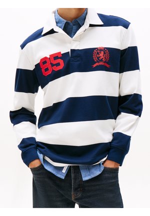 Homme portant un maillot de rugby bleu marine et blanc à rayures avec un "85" rouge et un emblème de blason, par-dessus une chemise bleue à boutons, les mains dans les poches d'un jean foncé.