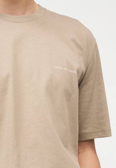 Μπεζ κοντομάνικο T-shirt με μικρό λευκό κείμενο "Tiger of Sweden" στο αριστερό στήθος, φορεμένο από έναν άνθρωπο μπροστά σε απλό φόντο.