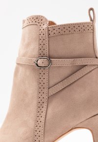 Bottine en suède beige avec des perforations, une sangle décorative avec une boucle en métal, et une surface texturée lisse.
