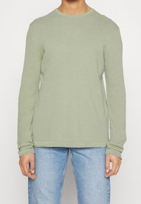 Pulls à col rond à manches longues en vert clair, fabriqué à partir d'un tissu tricoté texturé. Il présente une coupe décontractée et des poignets côtelés, associé à un jean bleu.