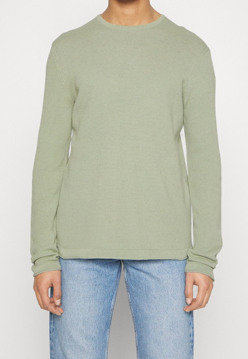 Pulls à col rond à manches longues en vert clair, fabriqué à partir d'un tissu tricoté texturé. Il présente une coupe décontractée et des poignets côtelés, associé à un jean bleu.