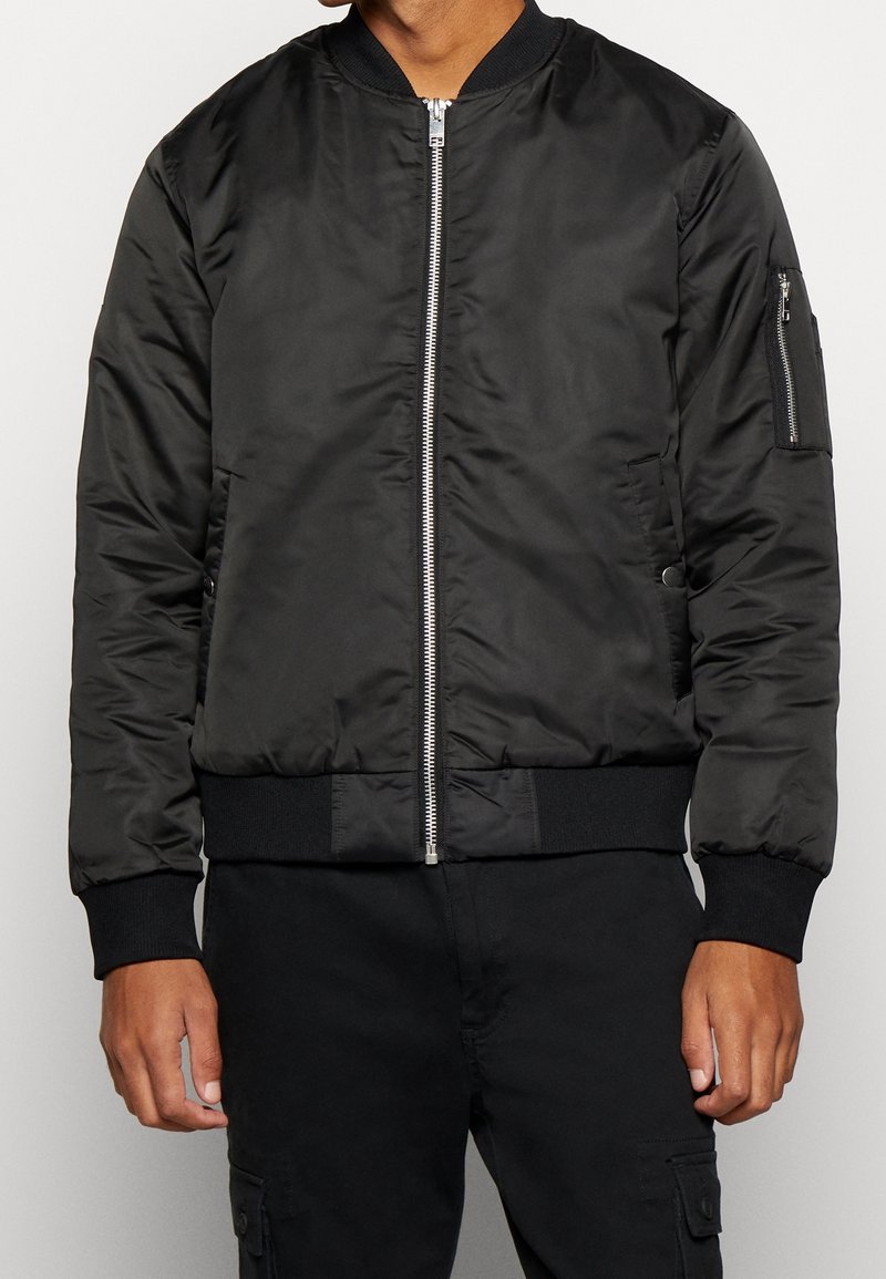Homme portant une veste bomber noire avec une fermeture éclair argentée sur le devant et une poche zippée sur la manche gauche.