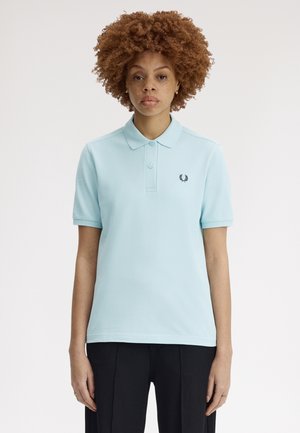 Donna con capelli ricci che indossa una polo azzurra chiara e pantaloni neri, in piedi su sfondo bianco, rivolta in avanti.