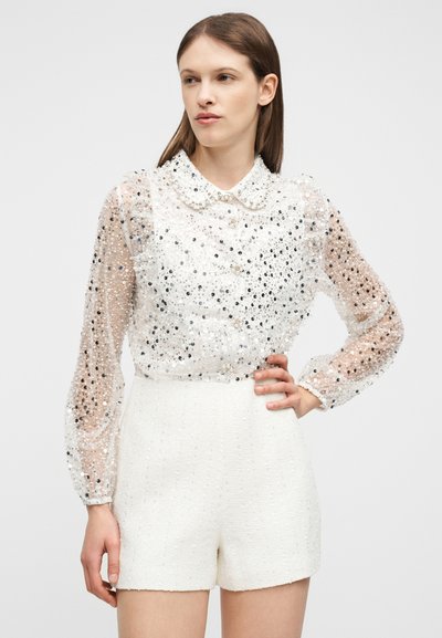 Femme portant un chemisier blanc transparent à manches longues orné de sequins et un short blanc texturé taille haute, debout avec une main sur la hanche.