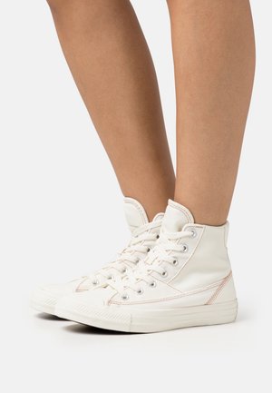 Gambe con sneaker alte bianche in tela con lacci, cuciture visibili e suole in gomma su uno sfondo bianco semplice.