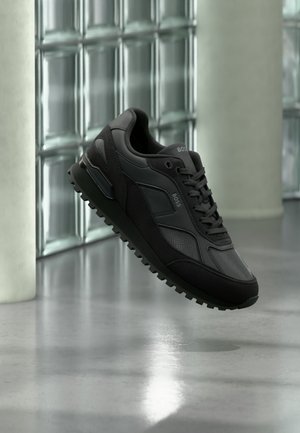 Czarny sportowy sneaker z teksturowanymi i gładkimi panelami, sznurowadłami i logo "BOSS", pokazany w powietrzu na tle ściany z bloków szklanych i polerowanej podłogi.