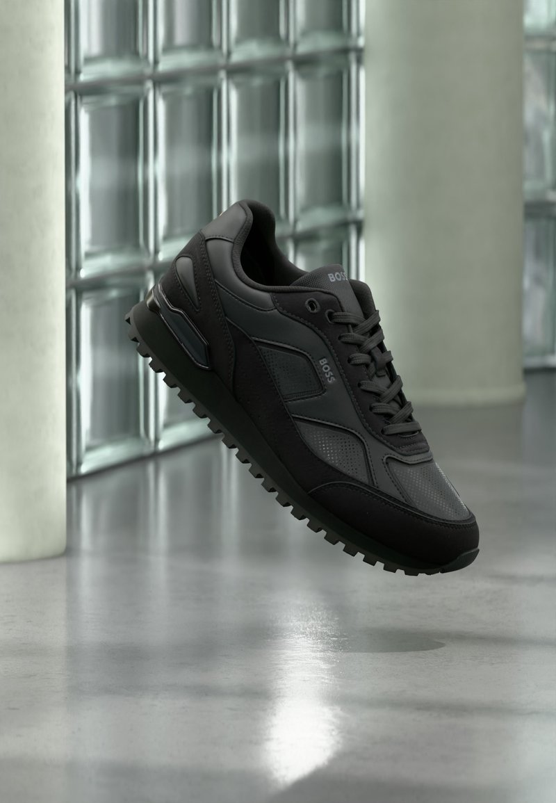Baskets de sport noires avec panneaux texturés et lisses, lacets et marquage "BOSS", montrées en suspension devant un mur en blocs de verre et un sol poli.