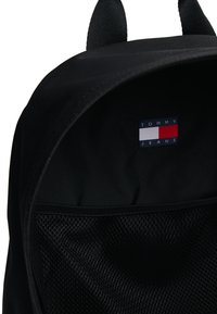 Svart ryggsäck av tyg med ett rektangulärt logotypmärke i rött, vitt och blått samt en nätficka för förvaring.