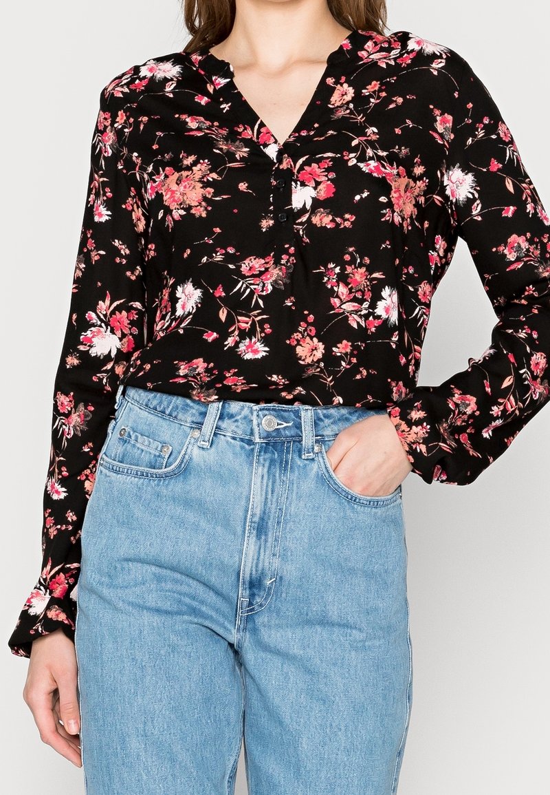 Femme portant un blouse noire à manches longues avec un imprimé floral rose, rentrée dans un jean en denim bleu clair taille haute, une main dans la poche.