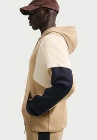 Sudadera con capucha en tonos beige y marrón claro, con mangas azul marino, que cuenta con un bolsillo tipo canguro y capucha. El diseño incluye bloques de colores contrastantes y tela texturizada.