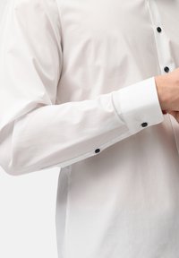 Camicia bianca con bottoni a lungo, caratterizzata da bottoni neri sui polsini, texture liscia e silhouette aderente.