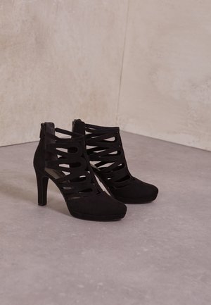 Czarne zamszowe buty na wysokim obcasie z otwartym noskiem, z wyciętymi paskami i zapięciem na zamek z tyłu. Gładka tekstura, zaokrąglona sylwetka.